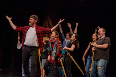 Crane in red checkered work shirt, white t-shirt, black jeans, backward baseball cap holds up his hands as he strides before a rabble of cheering people, all with wooden swords.